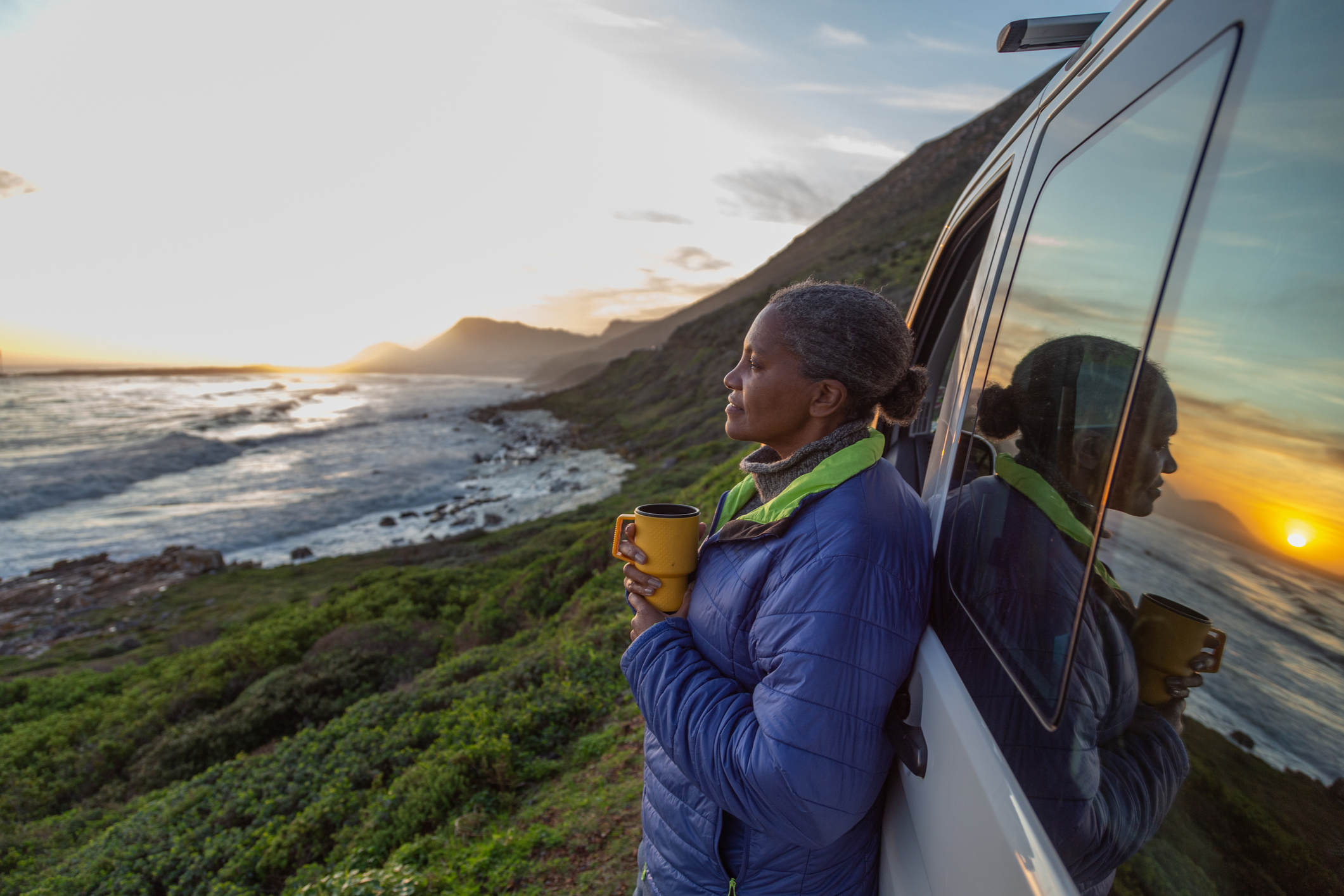 A person leans against a vehicle, holding a mug, gazing at an ocean and mountain view during sunset