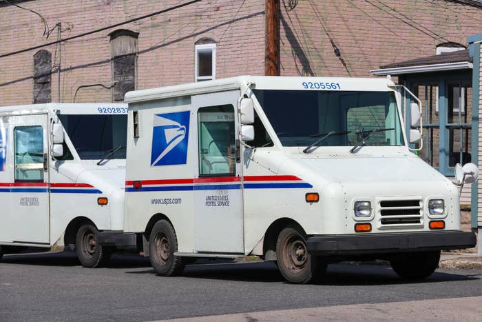 Two United States Postal Service (USPS) mail trucks are parked side by side on a street in front of brick buildings