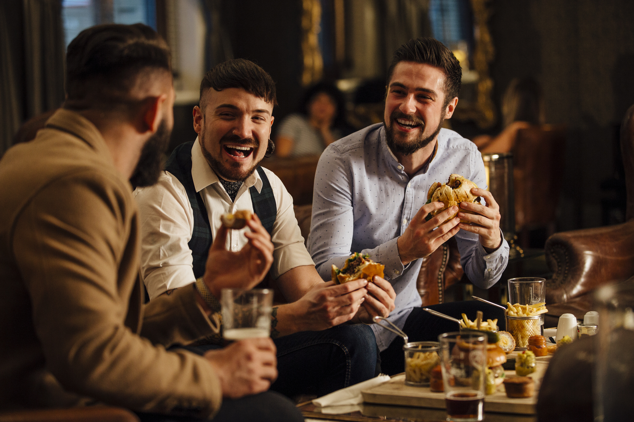 Three men, unknown identities, are sitting at a table laughing and eating burgers and fries in a casual setting