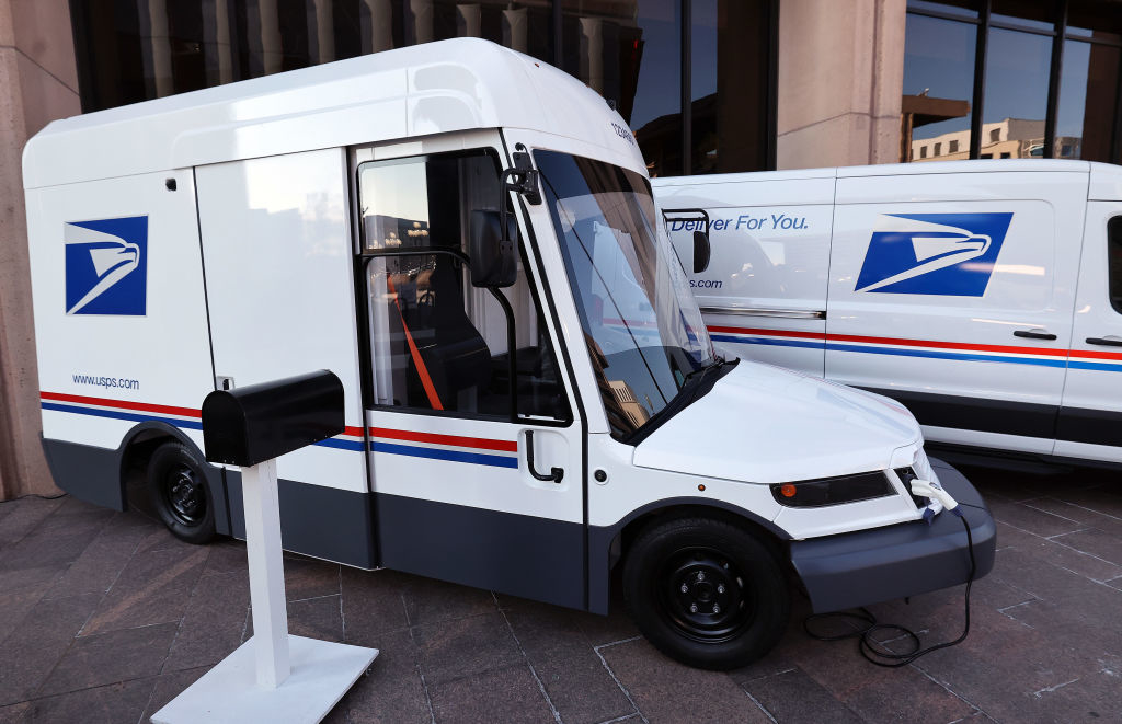 USPS electric delivery vehicle next to a mailbox, showcasing new eco-friendly fleet. Background: second USPS van and buildings