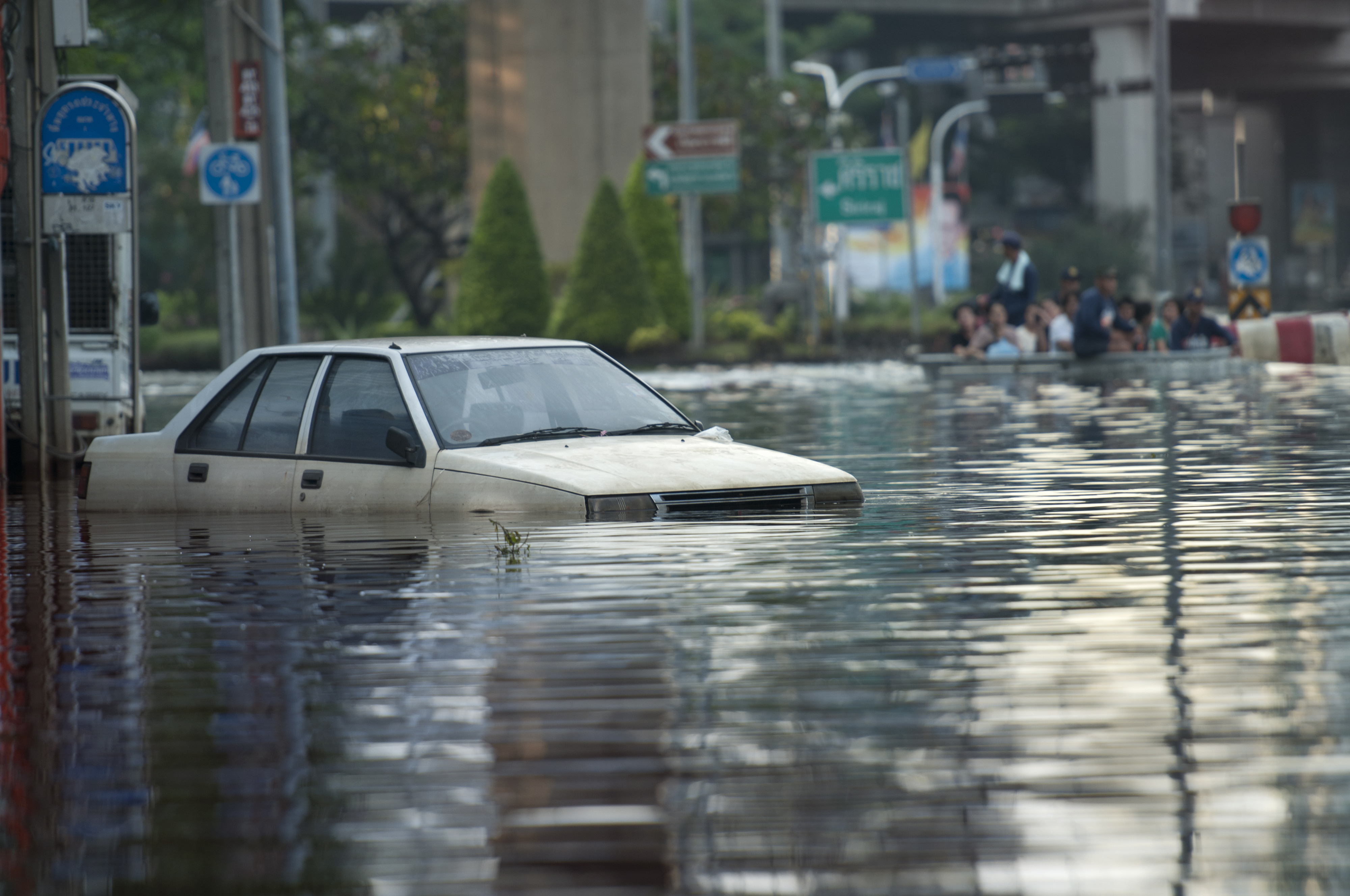 A white car is partially submerged in floodwaters on a street, with people visible in the background navigating the flooded area. Urban setting