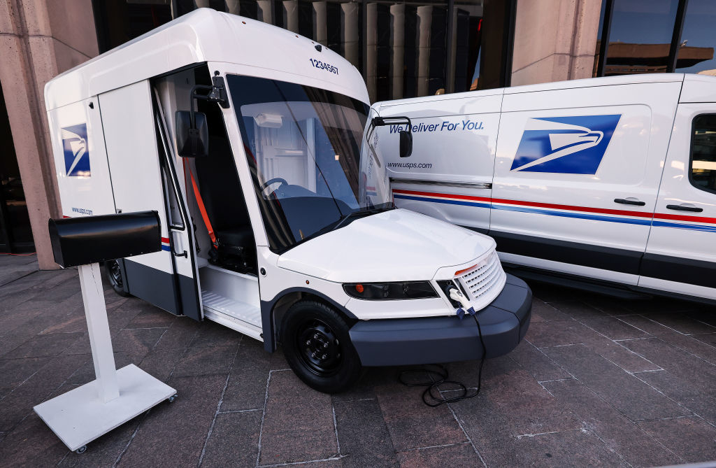 Two U.S. Postal Service delivery vehicles are parked, one with an electric charger connected, highlighting new electric mail trucks in the USPS fleet