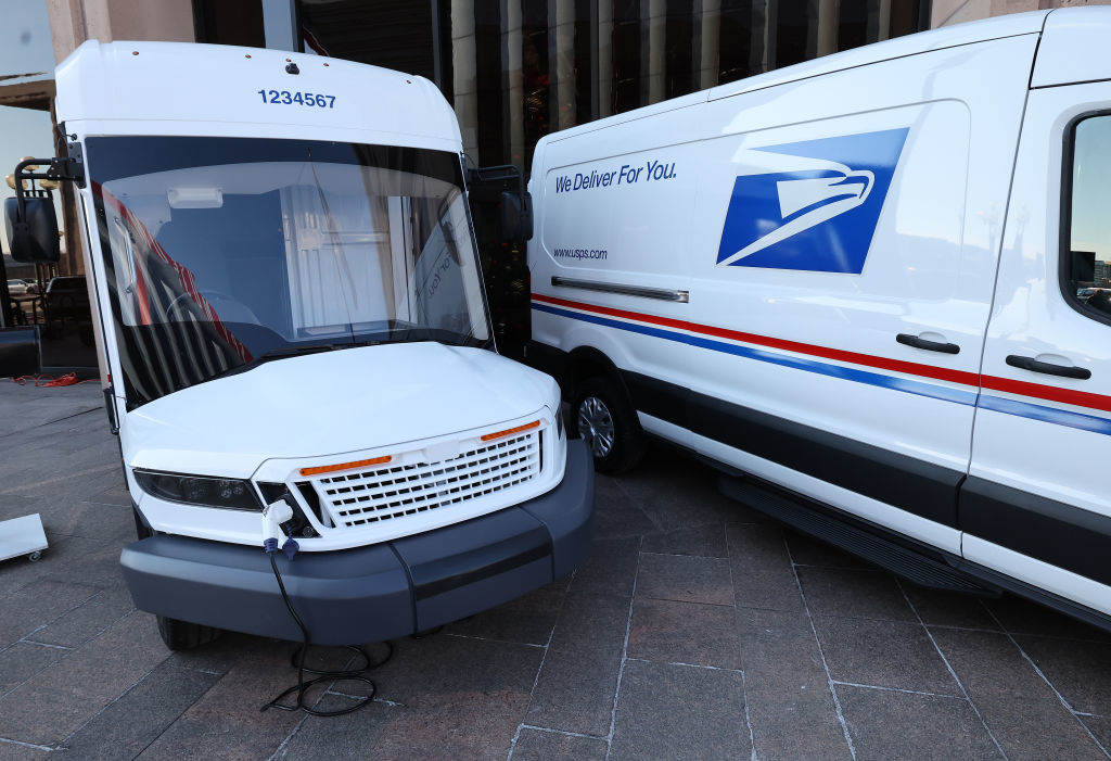 Two new USPS electric delivery vehicles parked side by side. One vehicle has &quot;We Deliver For You&quot; and the USPS logo on its side