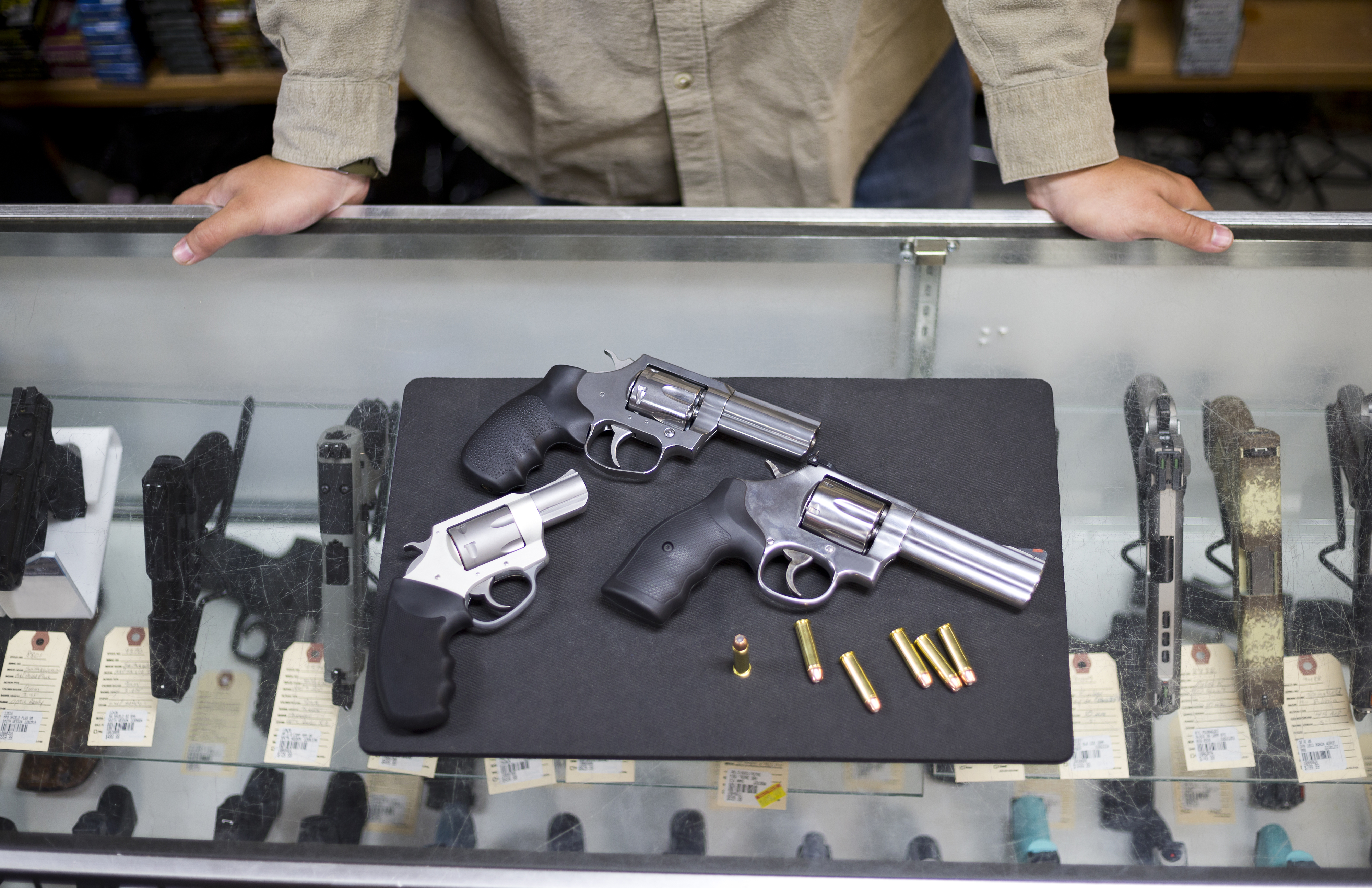 Three handguns and several bullets are displayed on a glass counter in a gun shop. A person stands behind the counter with hands resting on it