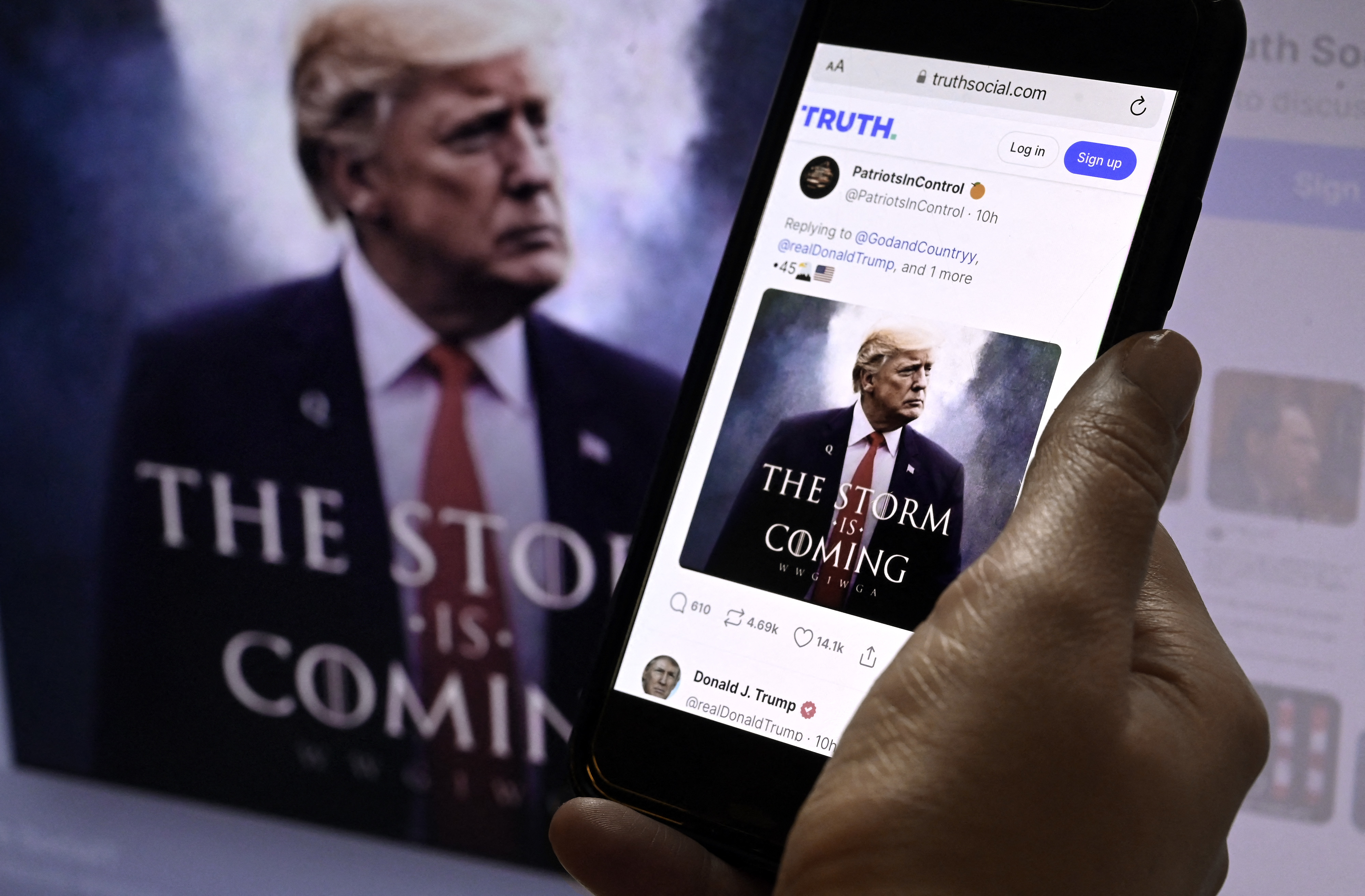 A hand holds a phone displaying a Truth Social post with an image of Donald J. Trump with the message &quot;The Storm Is Coming.&quot; Trump is also shown on a computer screen in the background