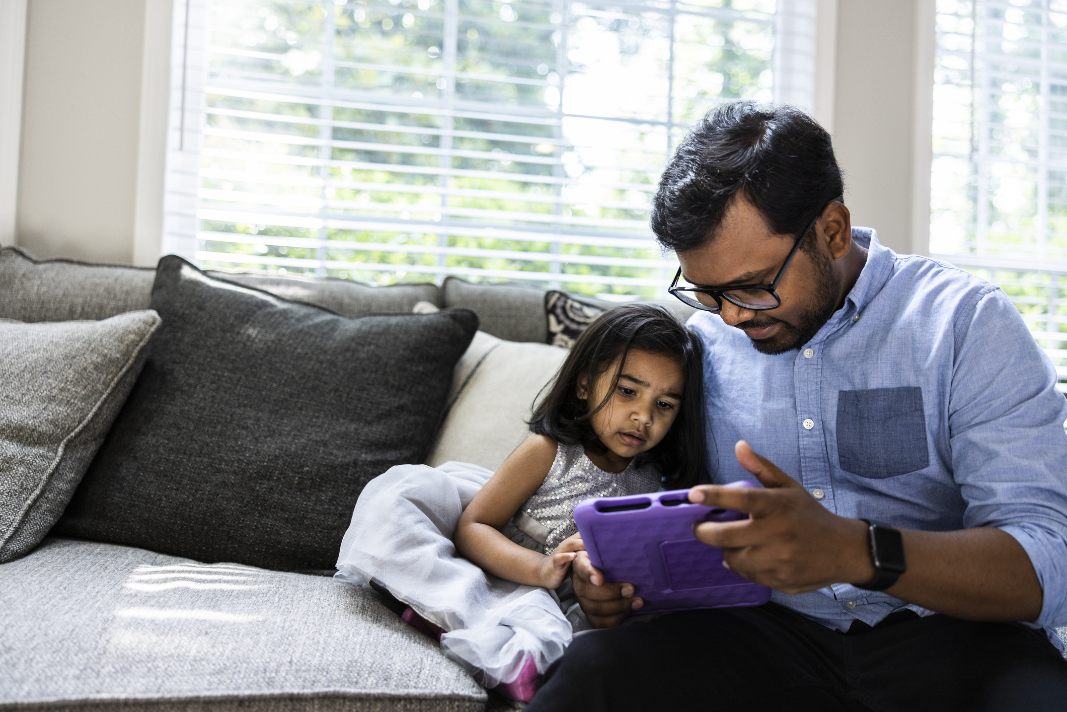 A man wearing glasses and a blue shirt sits on a couch with a young girl in a dress. They are looking at a tablet together