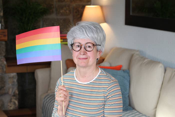 Older individual with grey hair and glasses, wearing a striped shirt and holding a rainbow flag inside a living room