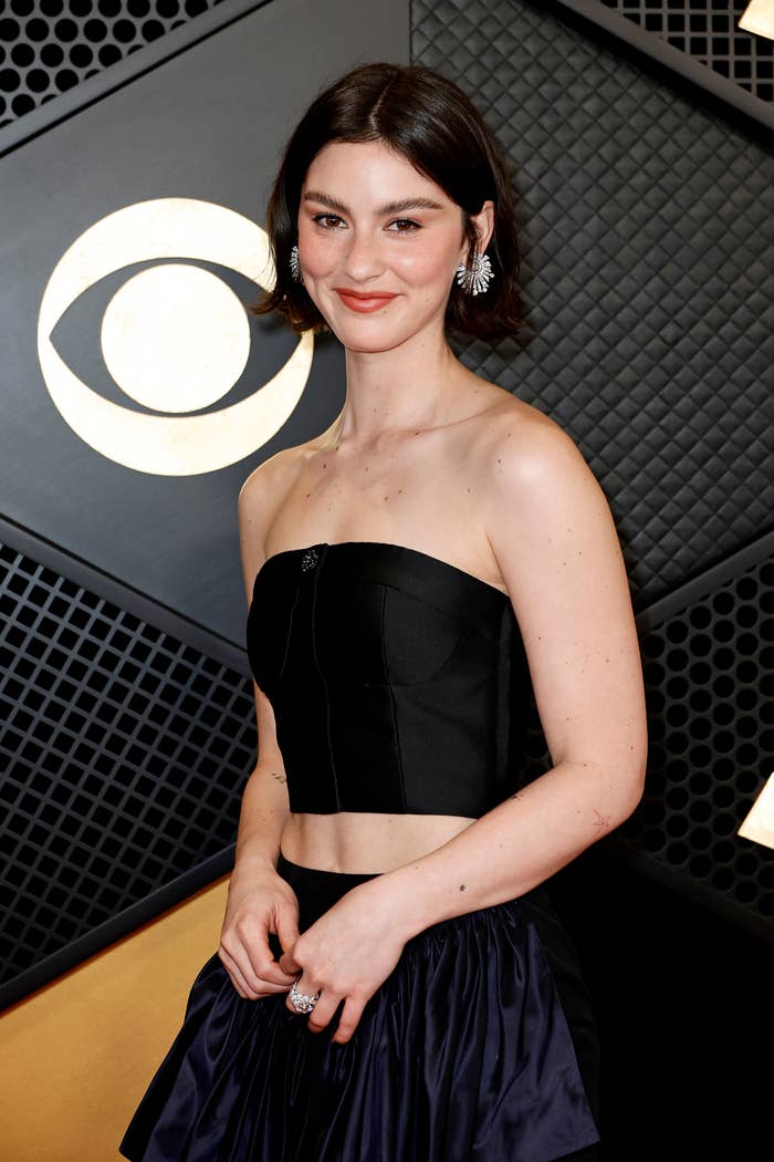A woman poses on a red carpet, wearing an elegant black strapless crop top and a matching skirt with statement earrings