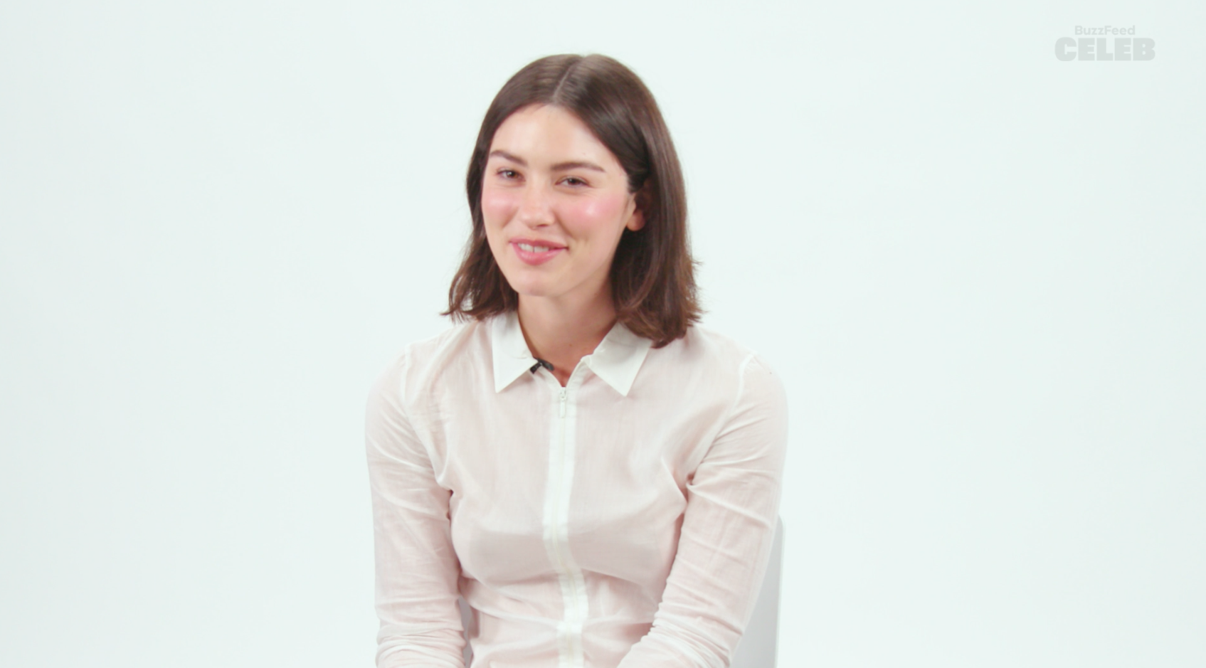 A woman with shoulder-length brown hair, wearing a long-sleeved, buttoned-up white shirt, sitting and smiling