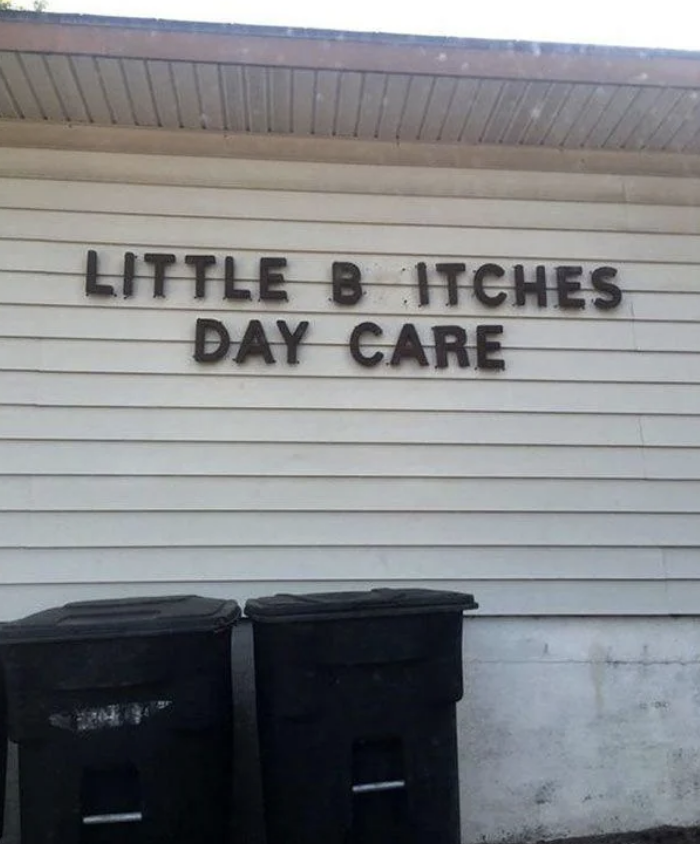 A daycare named &quot;Little Bitches Day Care&quot; has a sign on a building, with two trash bins placed below it