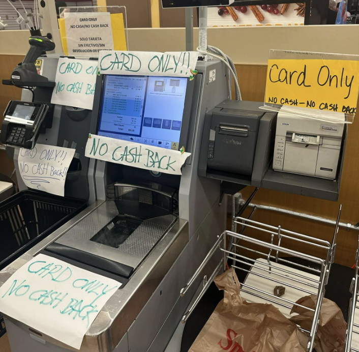 A self-checkout machine with multiple signs stating &quot;CARD ONLY!! NO CASH BACK.&quot; A shopping area is partially visible in the background