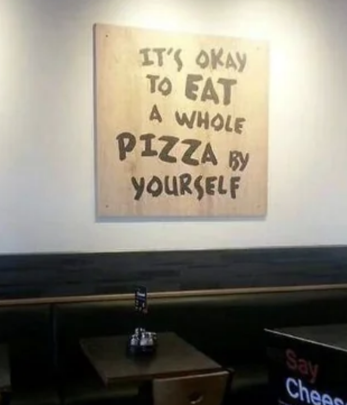 Wall sign reads, &quot;It&#x27;s okay to eat a whole pizza by yourself.&quot; Below the sign is a table with a small sign holder on it