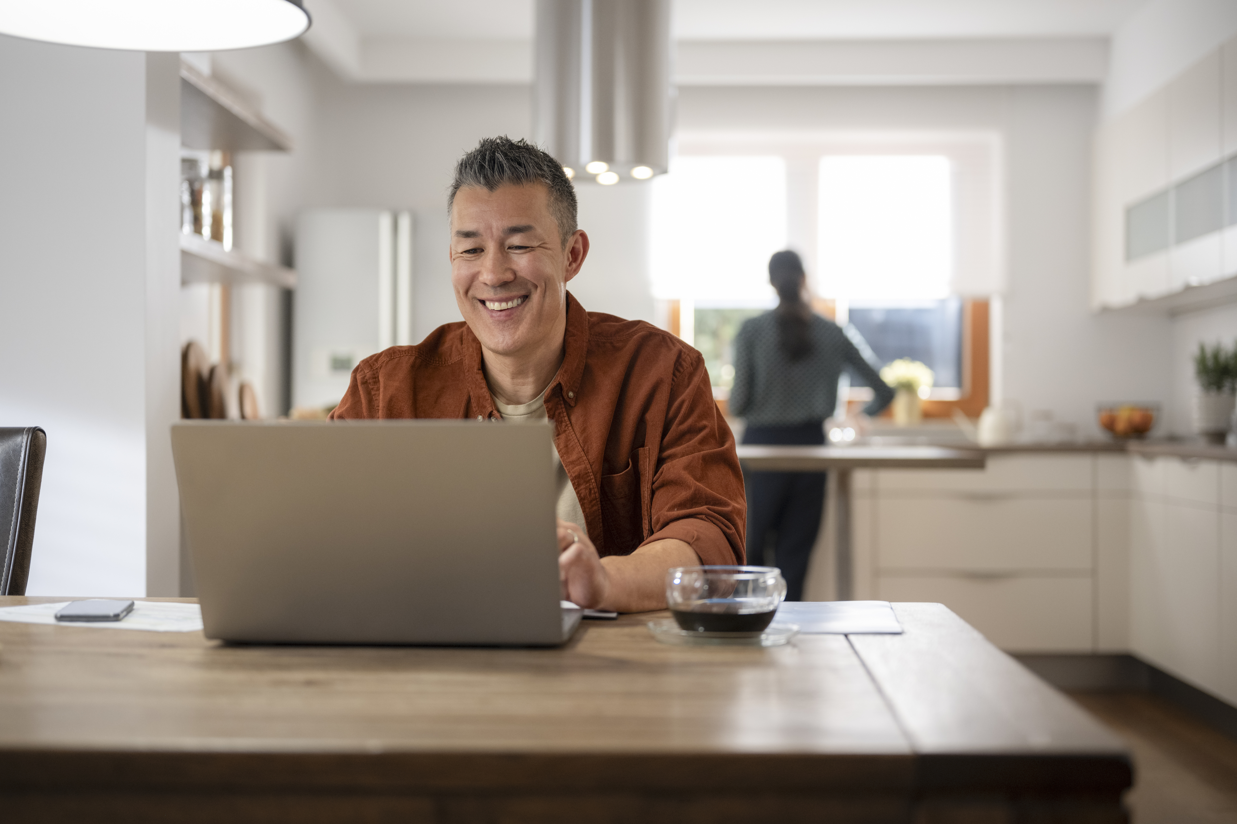 A man sits at a wooden table in a modern kitchen, smiling as he works on a laptop. Another person is blurred in the background near the kitchen area