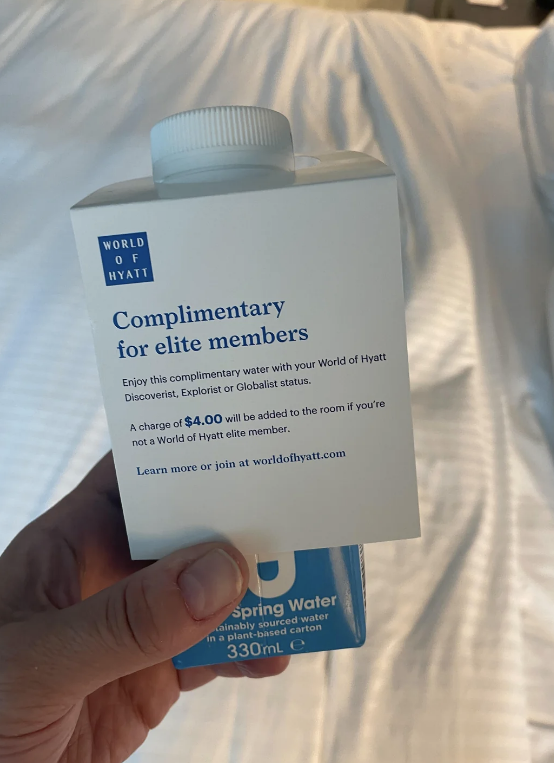 Hand holding a complimentary water carton by World of Hyatt, stating it&#x27;s for elite members with Discoverist, Explorist, or Globalist status or for a charge of $4.00