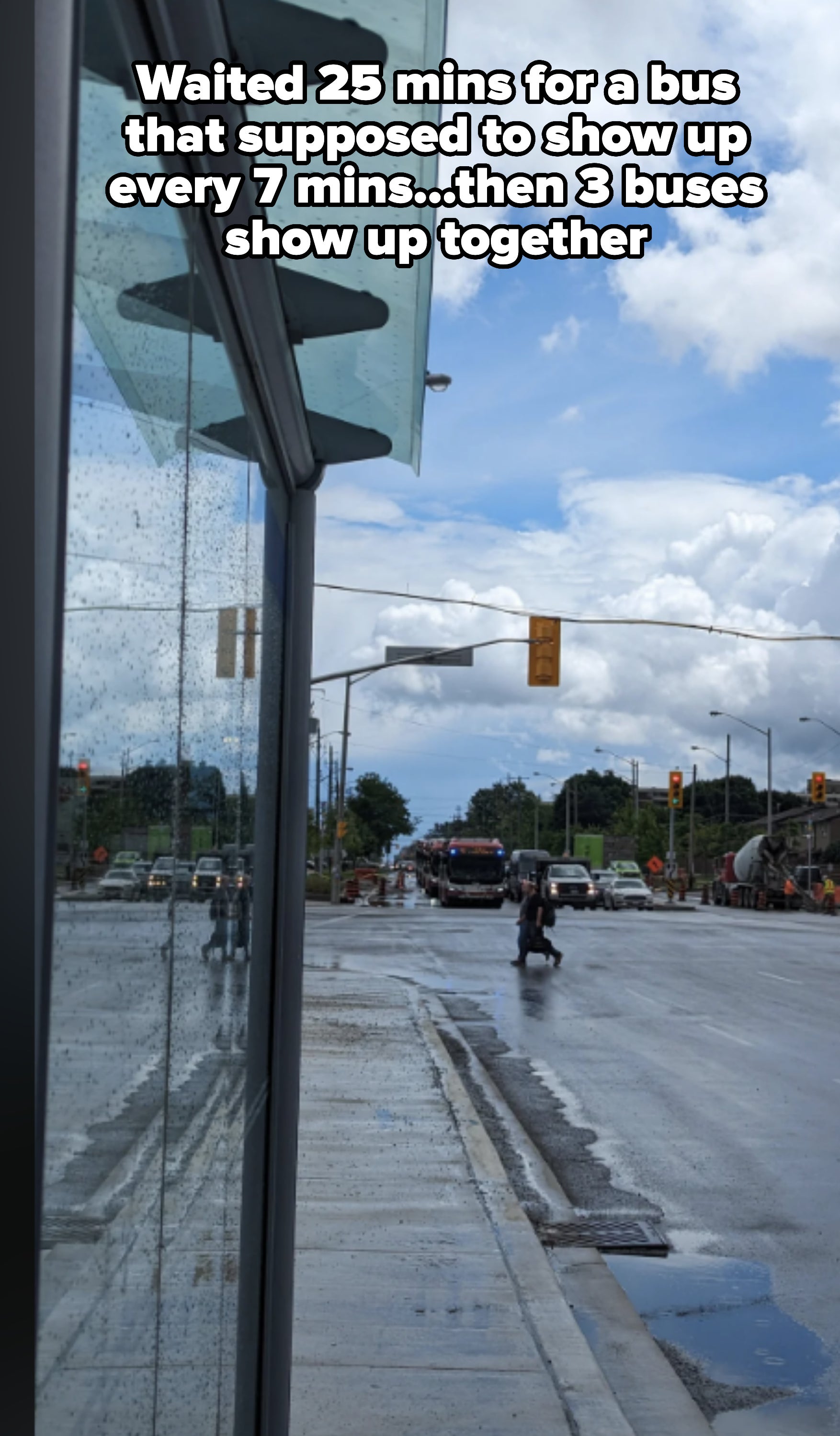 A rainy city street with traffic lights and vehicles in the distance. A person walks on the wet sidewalk near a glass building