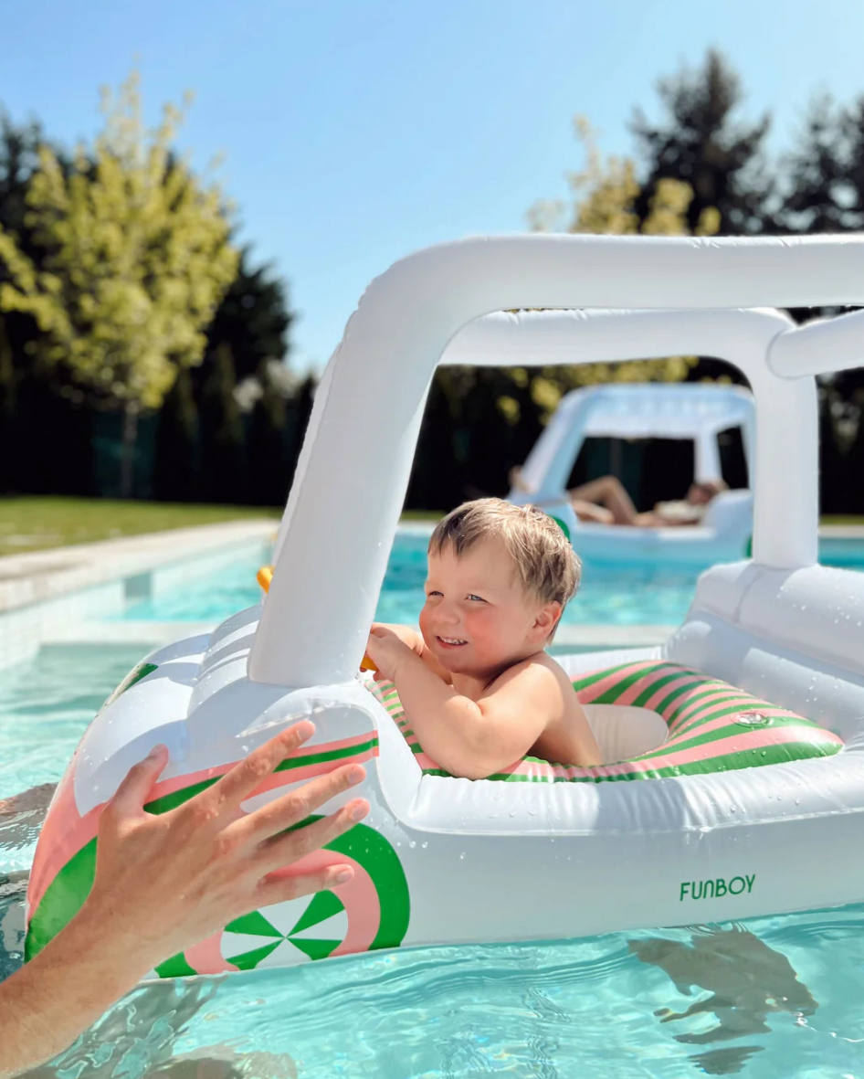 A child smiles while sitting in the golf cart pool float. An adult&#x27;s hand reaches out towards the child. The pool and trees are visible in the background