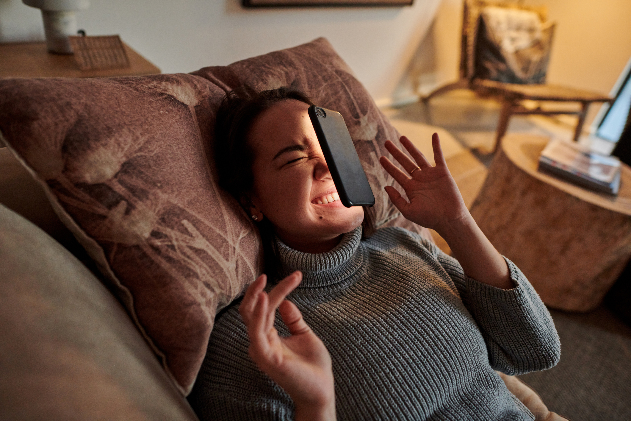 A woman lies on a couch with a textured pillow behind her, wincing and raising her hand as a smartphone falls onto her face