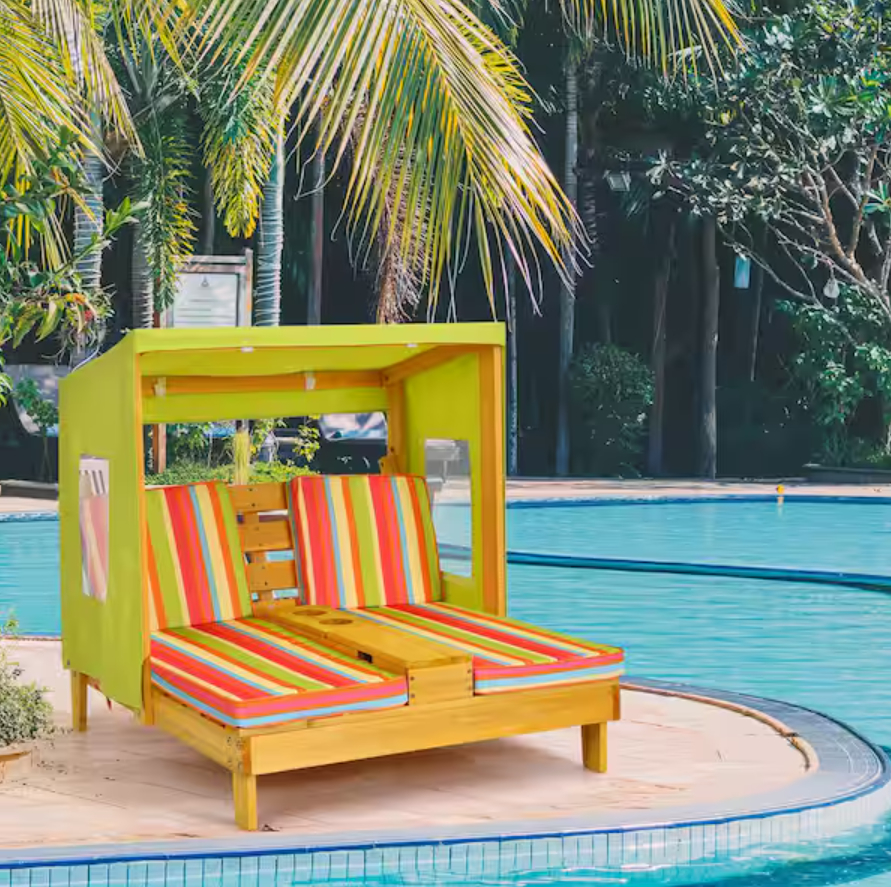 A poolside scene featuring a double sun lounger with a canopy, adorned with colorful striped cushions. Palm trees and greenery are in the background. No people are present