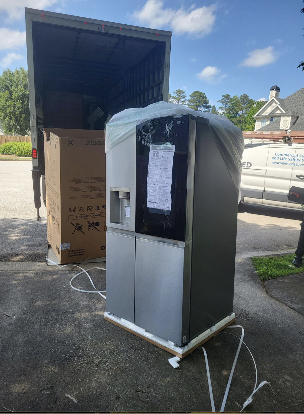 A large LG refrigerator, partially unwrapped with protective covering, stands in front of a truck. An unassembled box is seen in the background