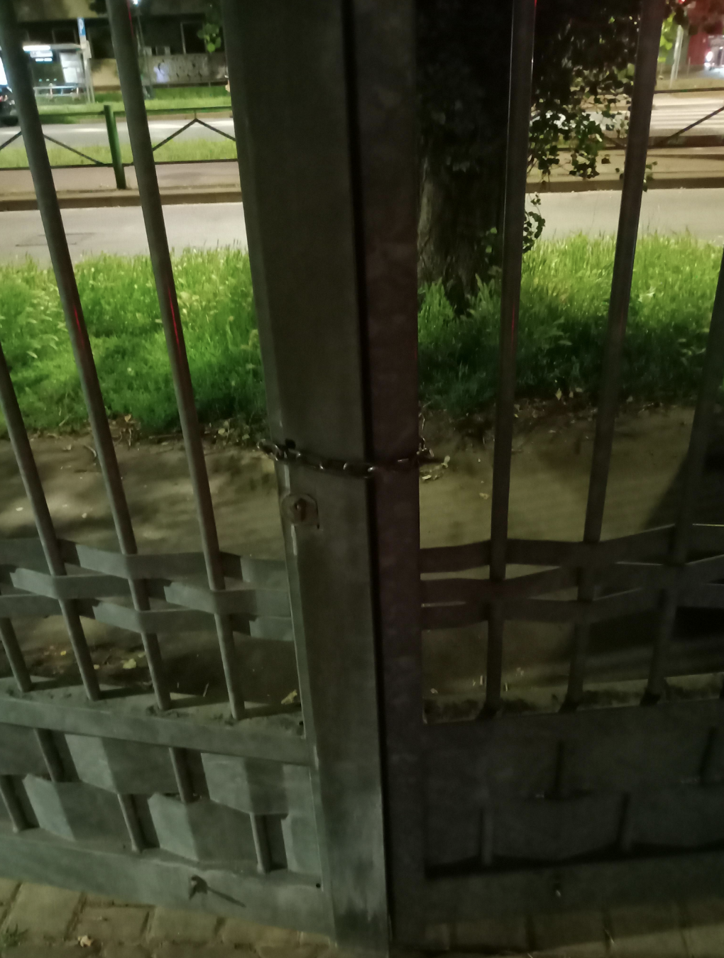 A metal gate chained and locked, preventing entry. Urban street and greenery visible in the background