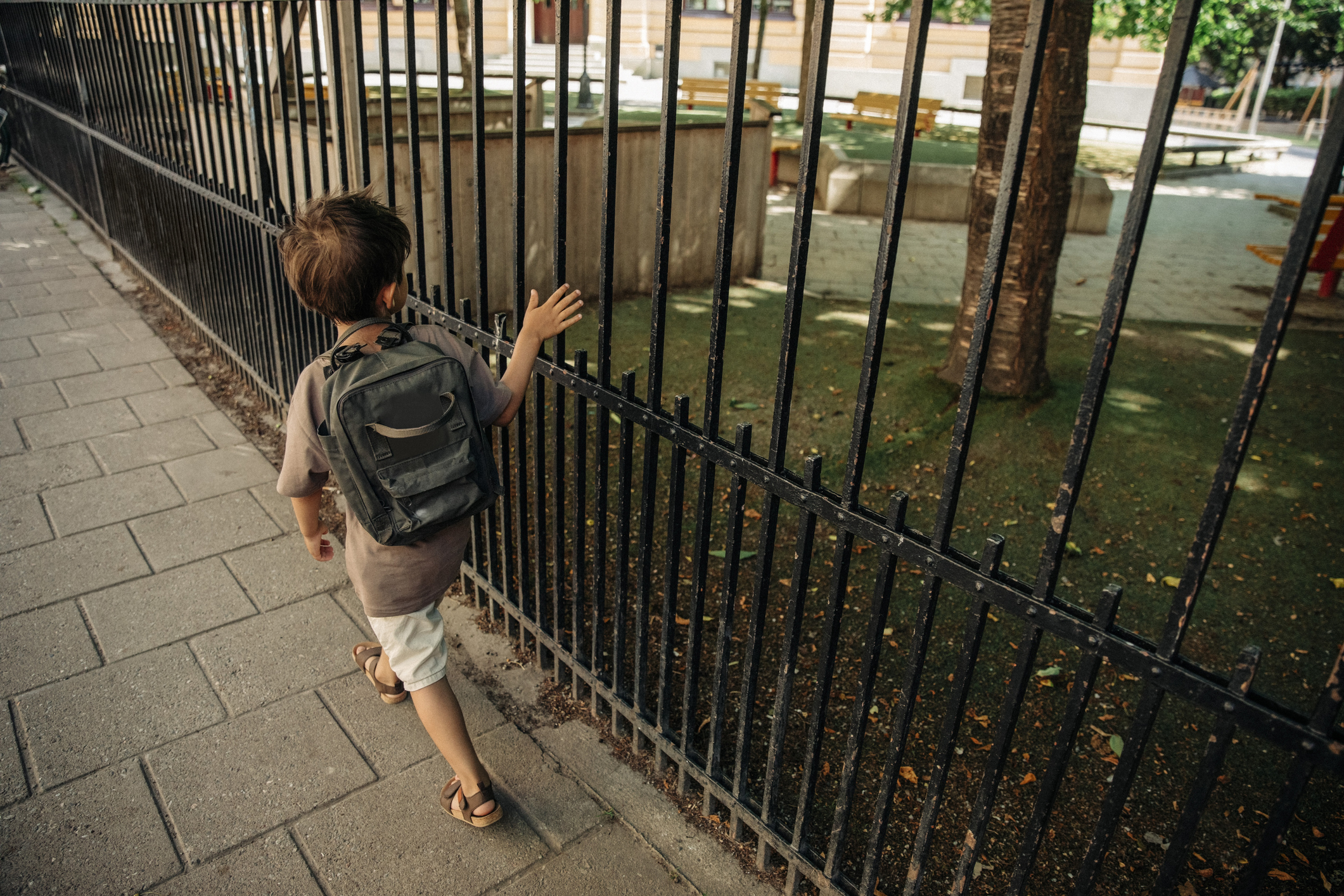 A young boy wearing a backpack walks along a fenced playground, touching the bars with his hand