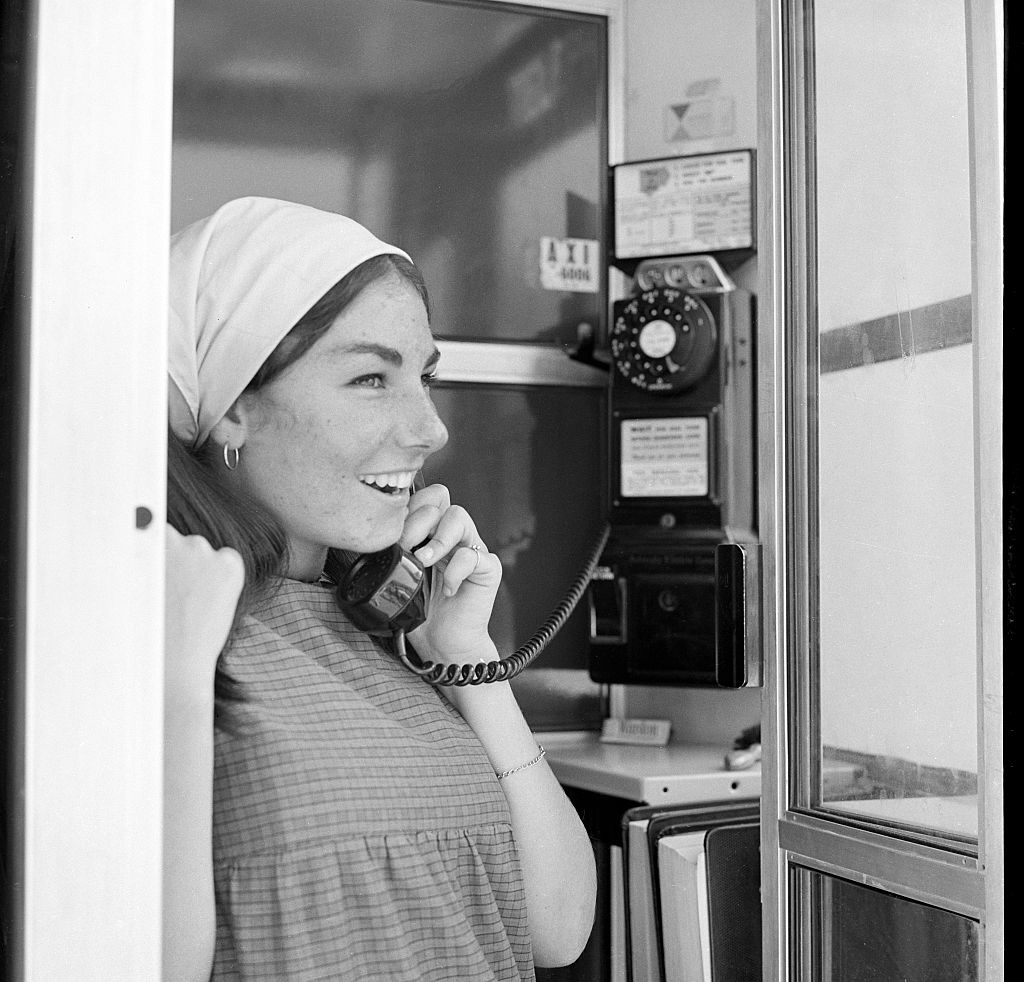 A woman standing in a phone booth, smiling and holding the receiver to her ear, wearing a headscarf and a checkered dress