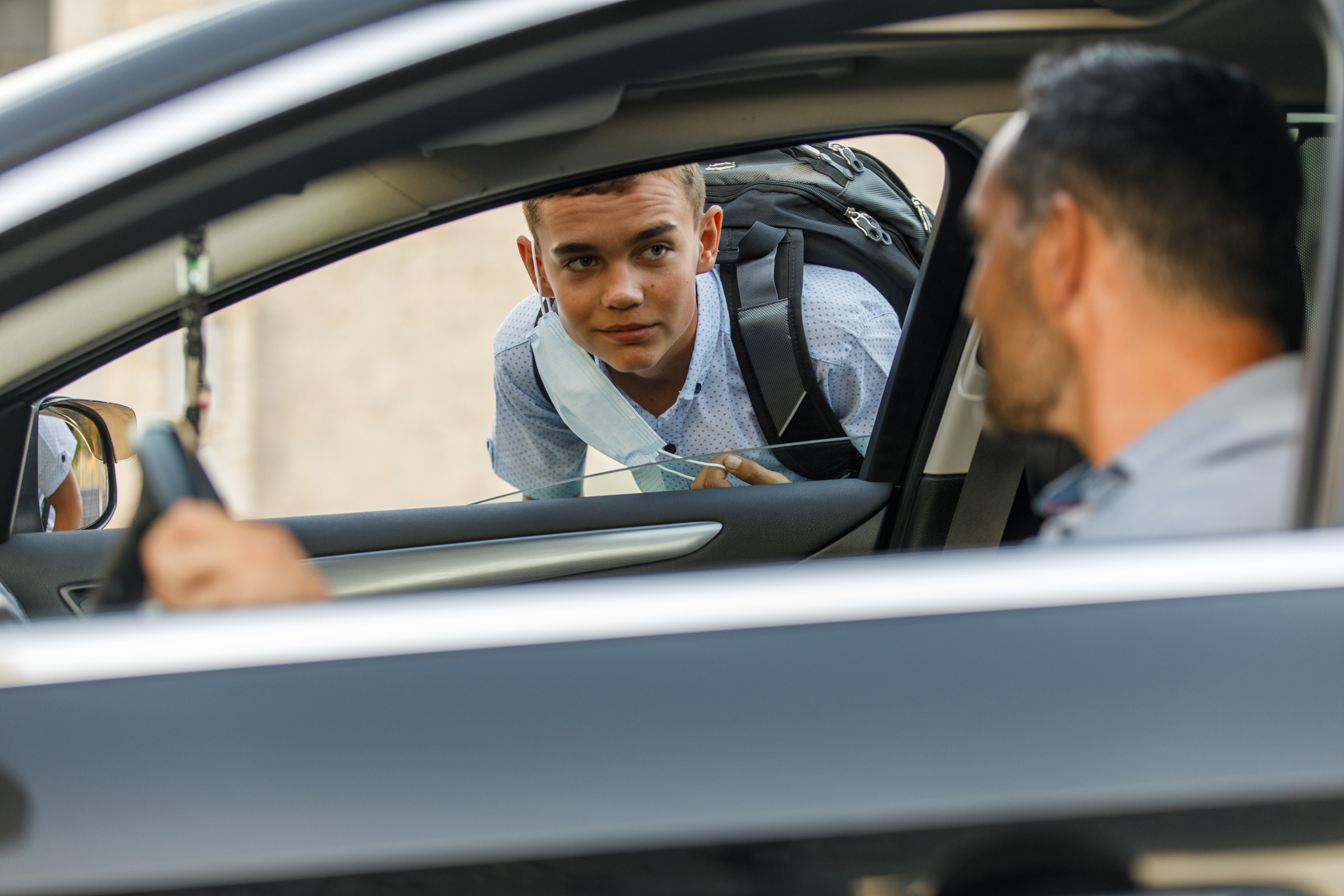 A teenage boy with a backpack talks to a man driving a car through the open passenger window