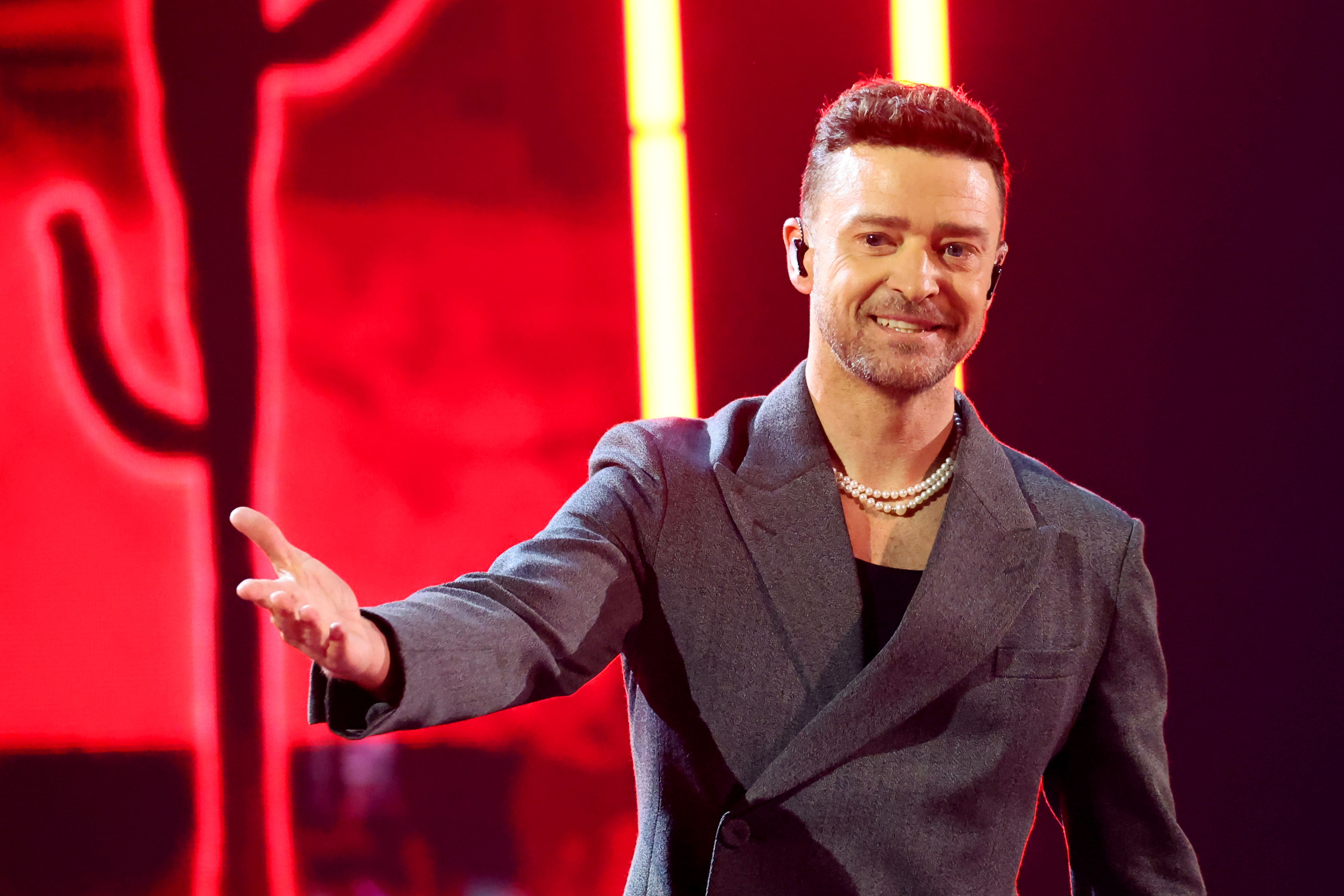 Justin Timberlake on stage, wearing a dark double-breasted blazer with a pearl necklace, smiling and extending his right hand