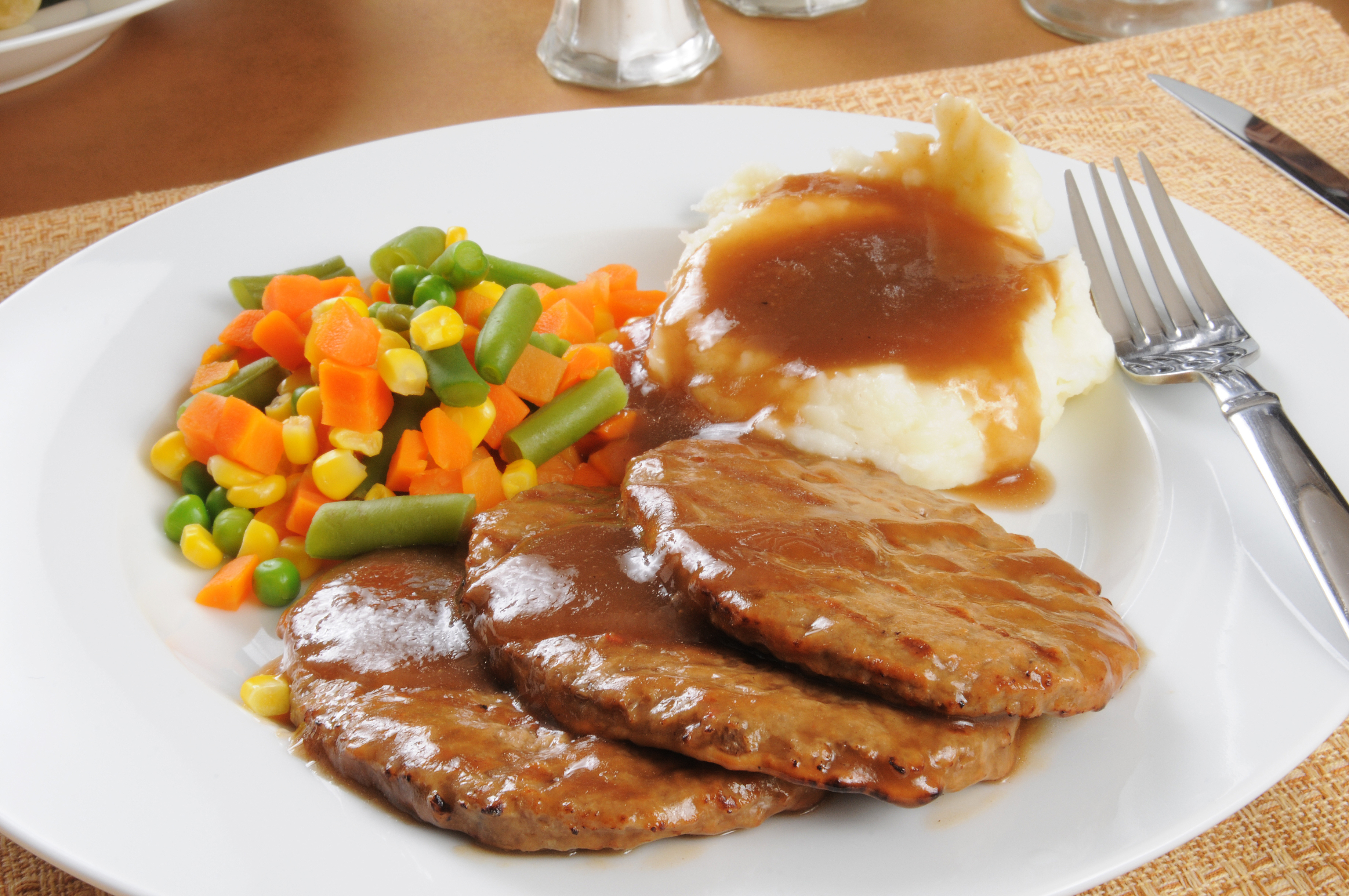 A plate of meatloaf topped with gravy, served with mashed potatoes and gravy, and a side of mixed vegetables (corn, peas, carrots, and green beans)