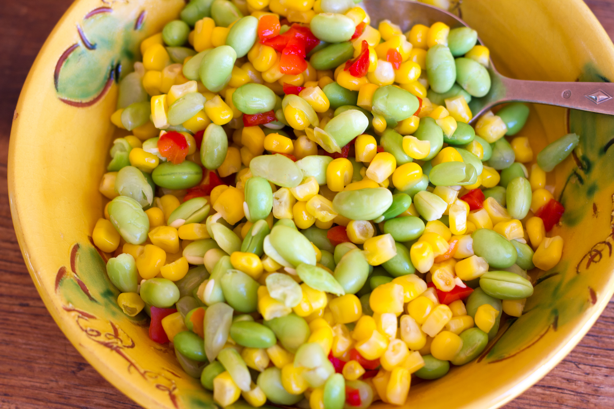 A close-up of a bowl filled with a mixed vegetable salad containing corn, edamame, and red bell peppers, with a serving spoon in the bowl