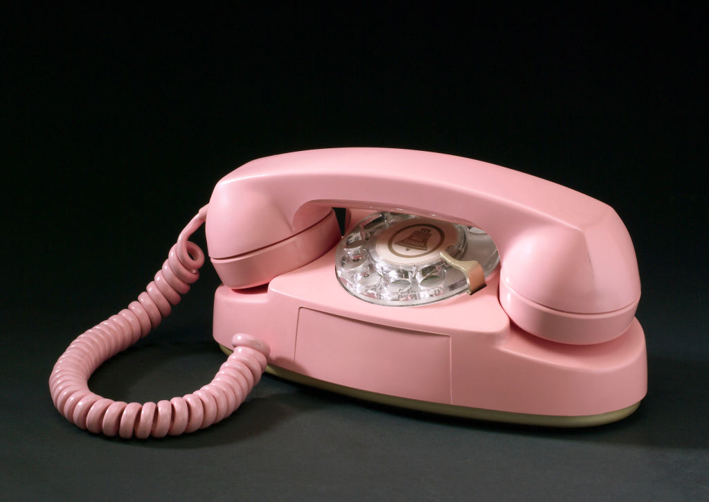 A pink rotary dial telephone with a coiled handset cord is displayed against a black background