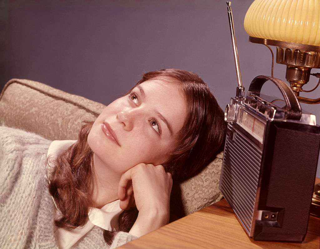 A young person with long hair, wearing a sweater and collared shirt, rests their head on a sofa arm while gazing thoughtfully. An old-fashioned radio sits nearby