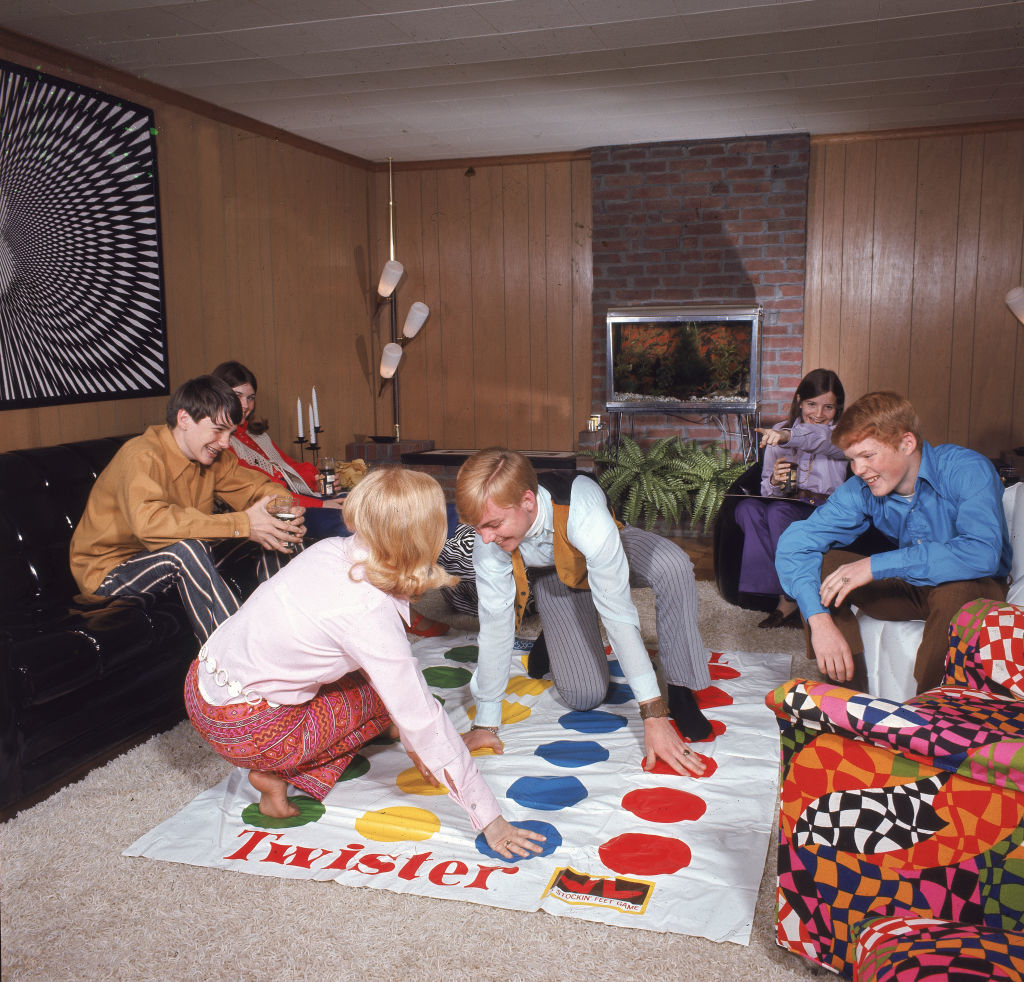 People playing a game of Twister in a 1970s-style living room with wood-paneled walls, a brick fireplace, and white shag carpet