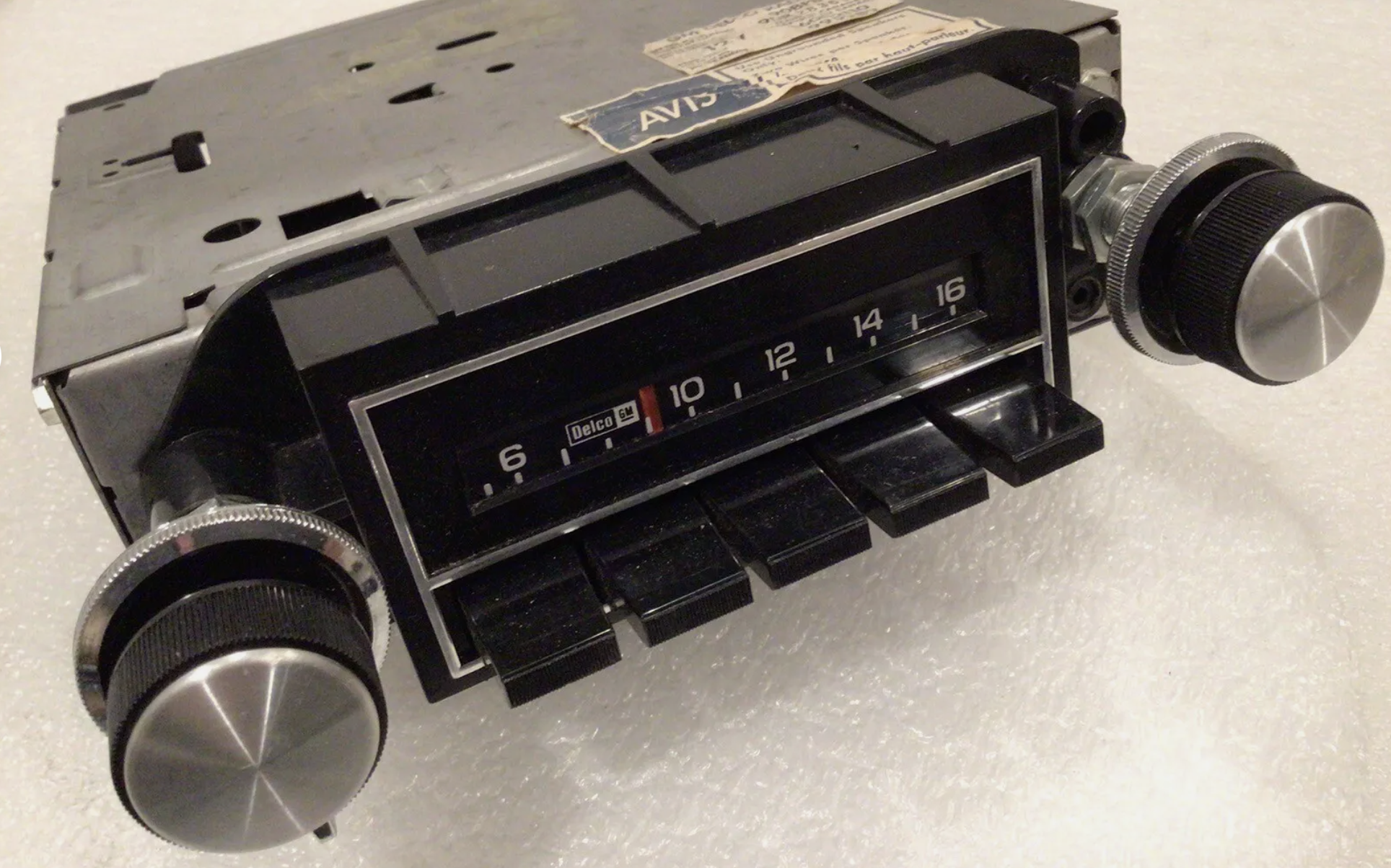 Vintage car radio with black knobs, a tuning dial, numbered scale from 6 to 16, and labeled &quot;Delco.&quot; The unit has visible wear and a partial sticker