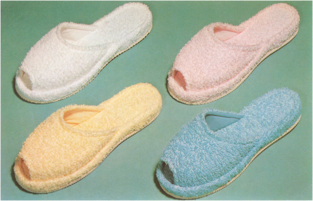 Four pairs of fuzzy, open-toe slippers in white, pink, yellow, and blue are arranged in a diagonal pattern on a flat surface