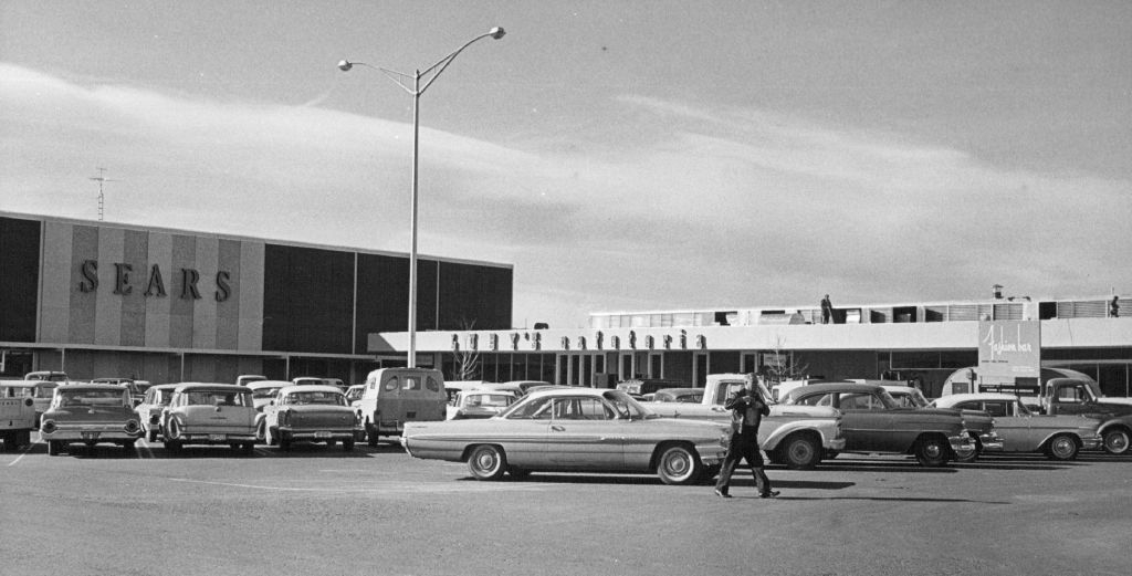 A vintage photo shows a busy parking lot outside a Sears store with numerous classic cars. A person is seen walking through the parking lot