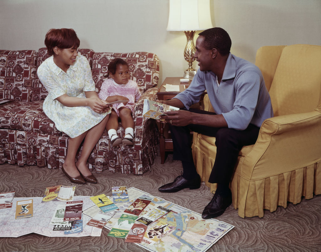 A woman, a man, and a child sit in a living room, surrounded by numerous travel brochures spread out on the floor. The man shows a brochure to the child