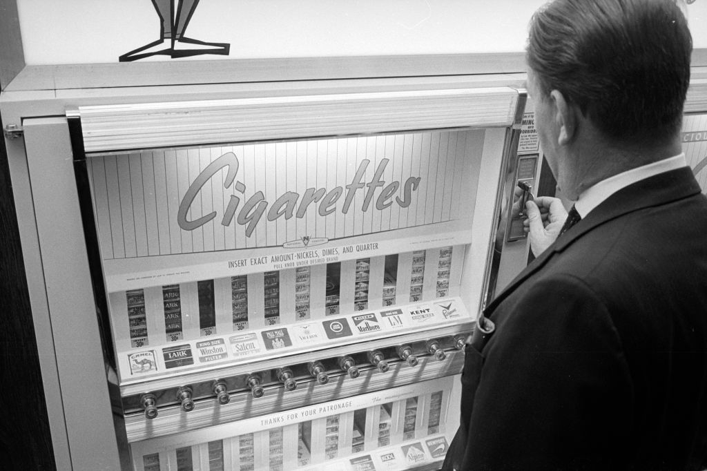 A man stands in front of a cigarette vending machine, preparing to make a selection. The machine displays various cigarette brands