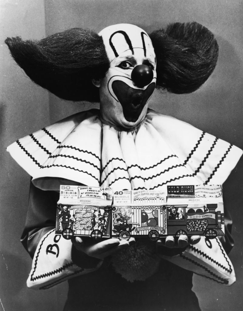 A vintage photo of Bozo the Clown holding two toy boxes labeled &quot;50 Tricks&quot; and &quot;40 Tricks&quot; with &quot;Gold Rush&quot; written on one. Bozo is in his iconic clown costume