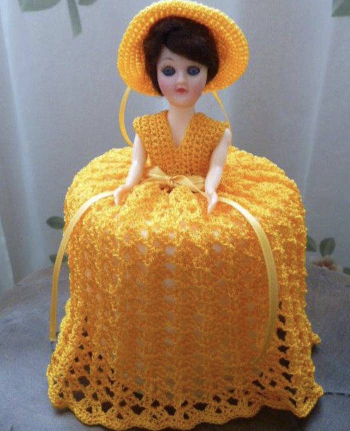 A vintage doll wearing a crocheted yellow dress and matching hat, seated on a surface, with a draped skirt