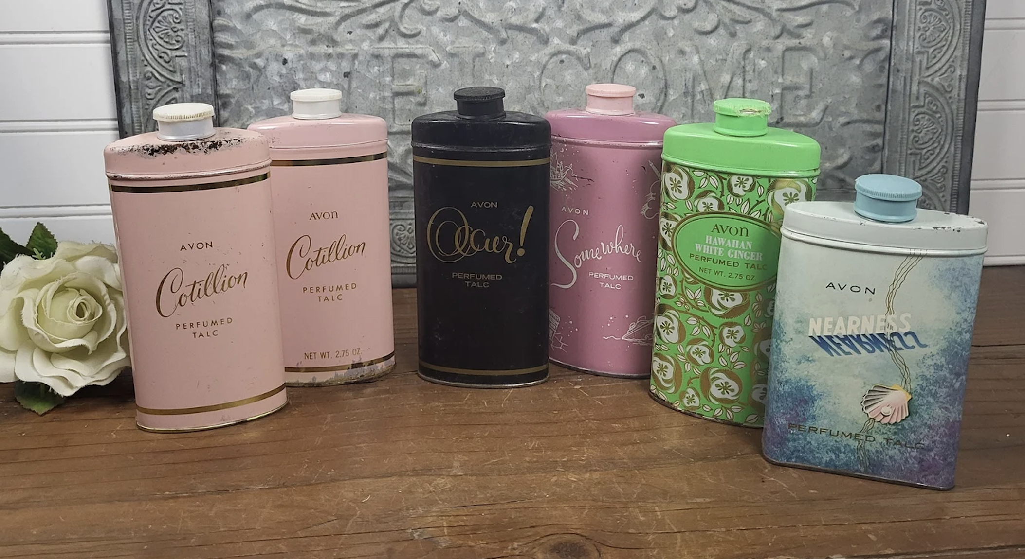 Six vintage Avon perfume talc tins are arranged in a row. The tins feature different labels and designs, with one tin depicting floral patterns