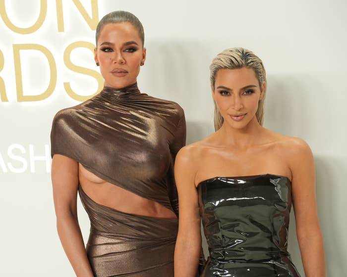 Kim Kardashian in a strapless dress, stands beside Khloé Kardashian who is wearing a metallic dress at an awards event with an asymmetrical cutout at the midriff