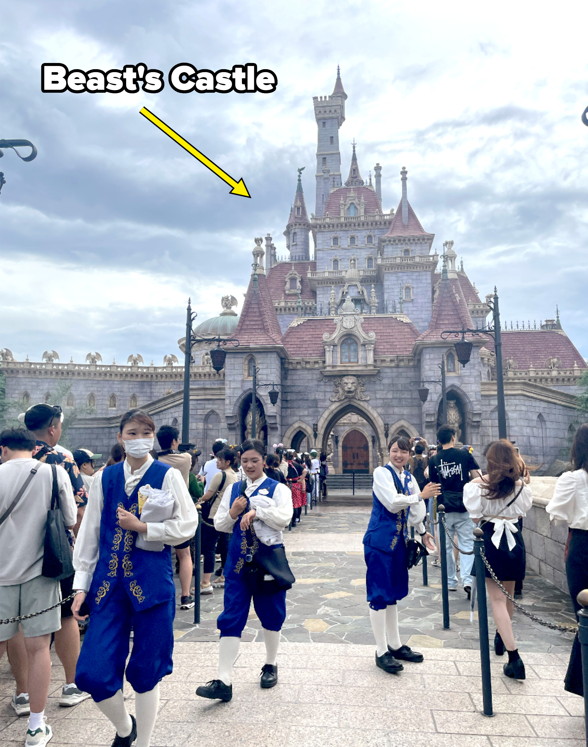 People dressed as Disney cast members in themed uniforms stand in front of the Beast&#x27;s Castle at Tokyo Disneyland, surrounded by visitors