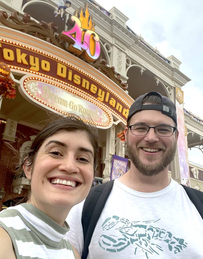 Two people smiling outside Tokyo Disneyland, celebrating its 40th anniversary. The park entrance reads &quot;Tokyo Disneyland&quot; and &quot;Dream Go Round.&quot;