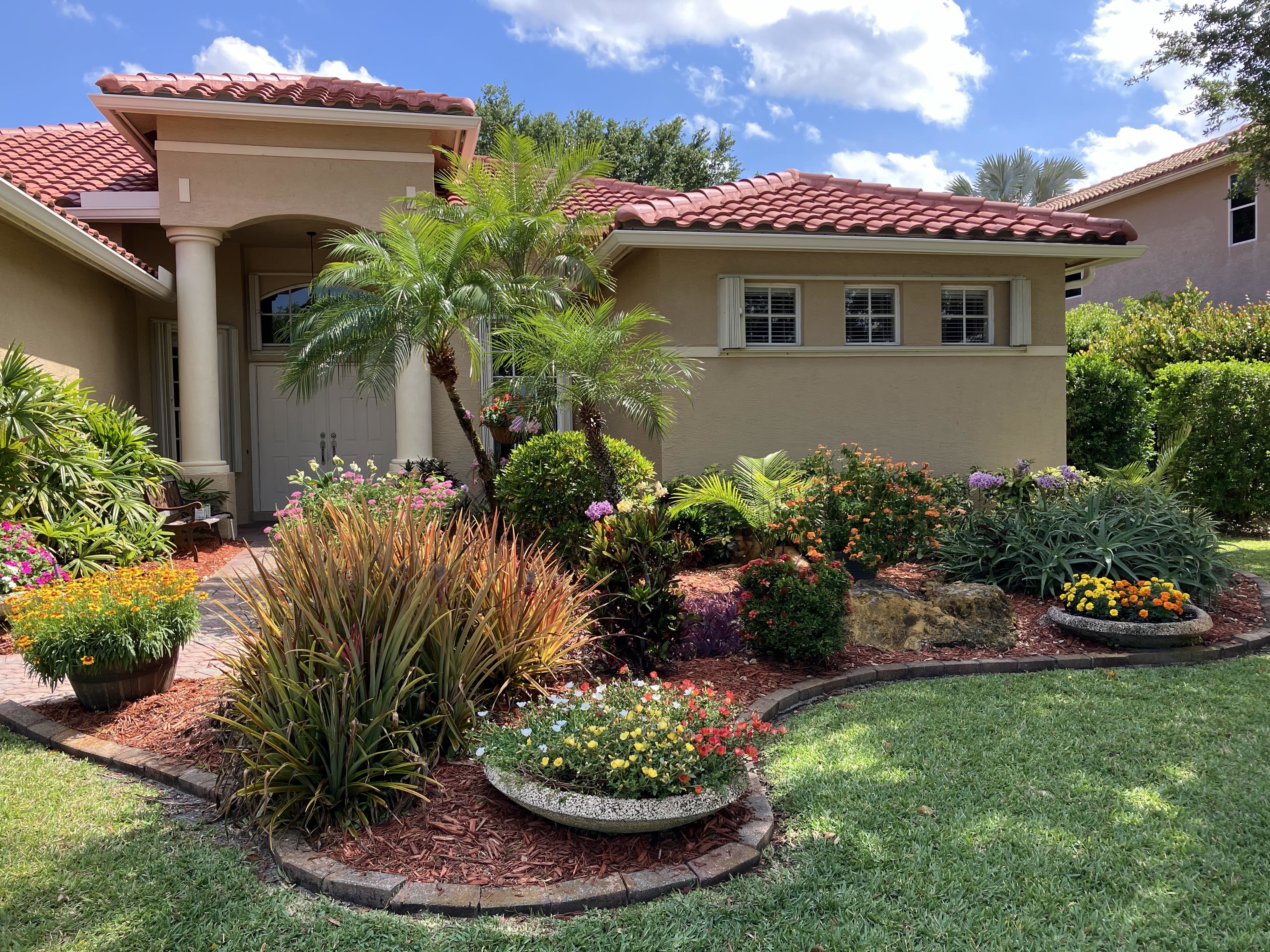 A well-maintained front yard of a house with a variety of flowering plants, shrubs, and a small palm tree. House has beige walls and a red-tiled roof