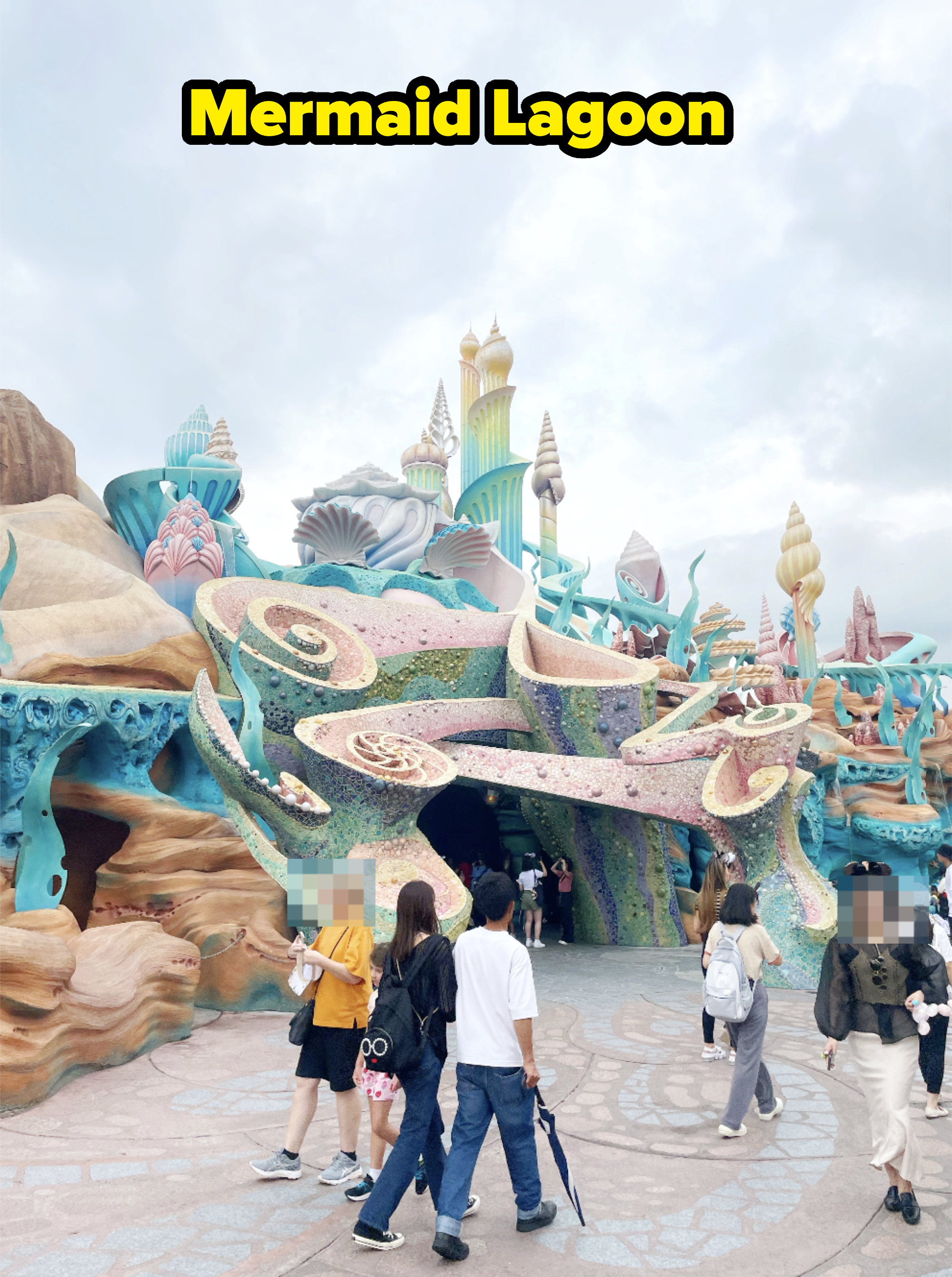 People walk around an imaginative, fantasy-themed Disney amusement park area with whimsical architecture, resembling undersea coral and seashell structures