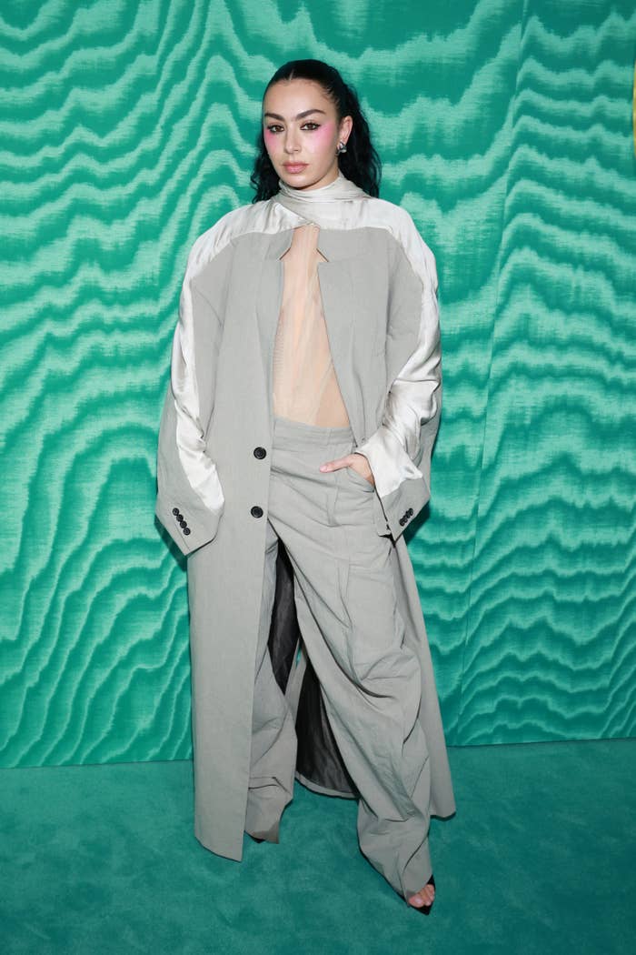 Charli xcx poses in a loose-fitting gray suit with a sheer top against a textured green backdrop