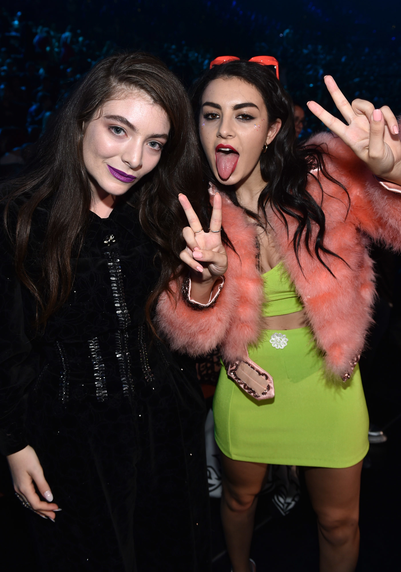 Lorde and Charli xcx posing together at an event. Lorde is wearing a black dress, and Charli xcx is wearing a green dress with a pink fur coat. Both are giving peace signs