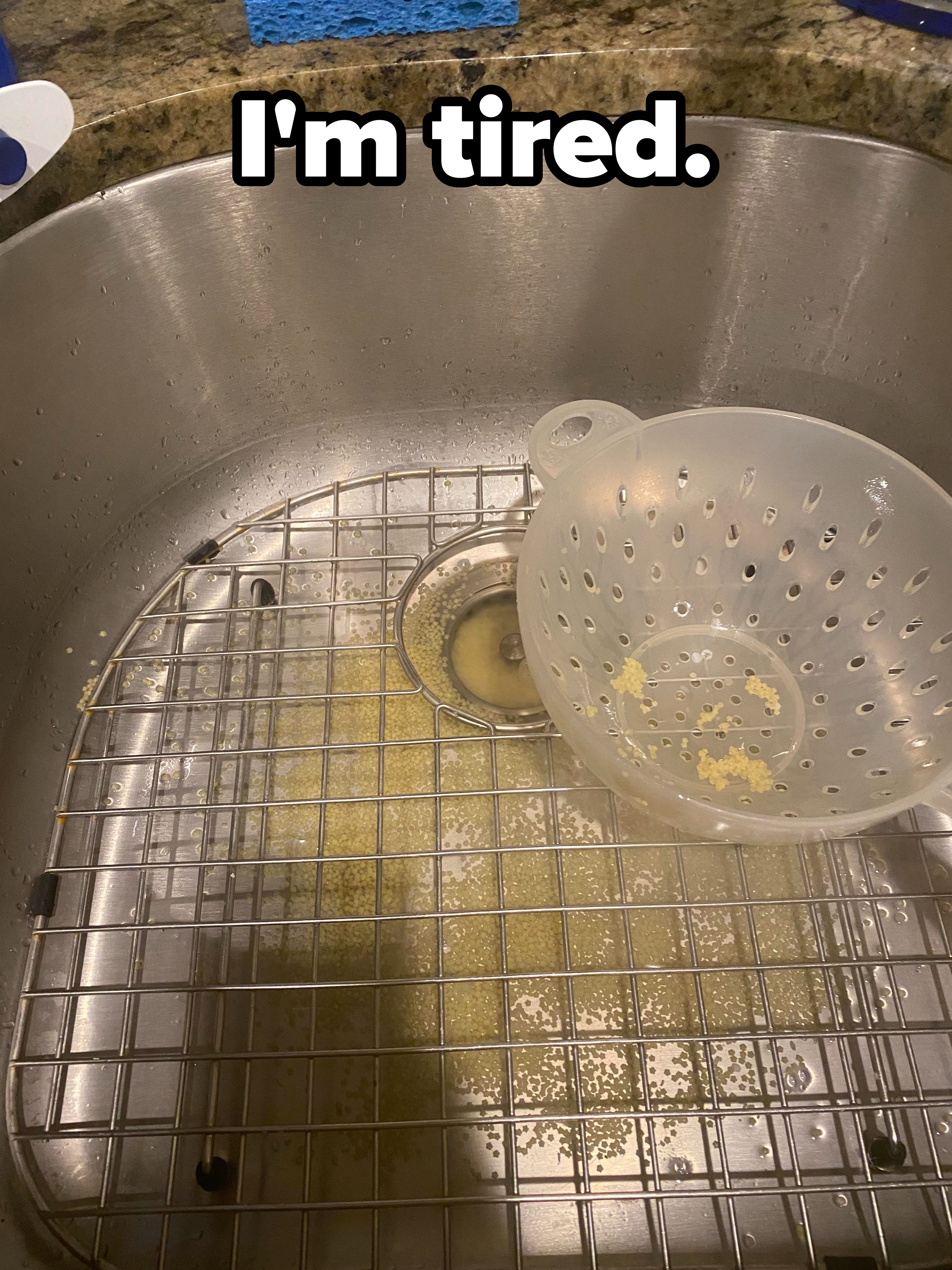 Colander with small bits of food residue is placed in a kitchen sink with some food remnants in the sink strainer