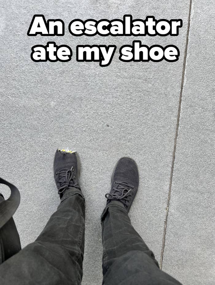 Person wearing black sneakers with one sneaker&#x27;s toe area visibly worn out, standing on a concrete surface. Only their legs and feet are visible